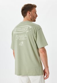 Camiseta de algodón verde claro con mangas cortas, que presenta una impresión blanca en la parte posterior. El texto incluye frases motivacionales y diseños gráficos simples.