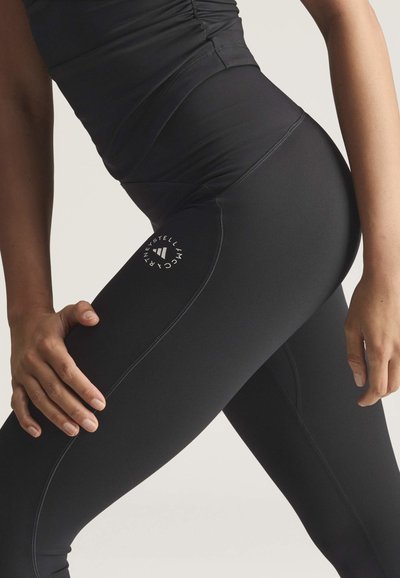 Close-up af en person iført sorte, højtaljede leggings med et hvidt, rundt logo på låret, hænderne hvilende på benene.