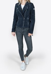 Woman wearing dark blue suede zip-up jacket, grey skinny jeans, and white sneakers, standing with one foot slightly forward.