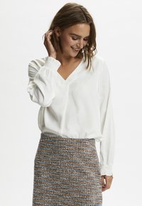 White, long-sleeve blouse with a V-neck, featuring gathered cuffs. Paired with a textured, multicolored skirt in woven patterns.