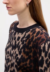 Pull en imprimé léopard avec un col noir rond, présentant des tons chaleureux de marron et de noir, fabriqué en tissu doux et texturé. Boucles d'oreilles en argent visibles.
