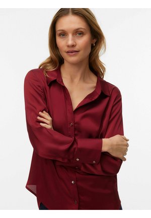 Femme à la peau claire et aux cheveux blonds porte une chemise en satin rouge foncé boutonnée à manches longues, se tenant les bras croisés devant un fond uni.