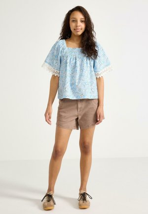 Nuori henkilö seisoo, pukeutuneena vaaleansiniseen kuvioituun puseroon, jossa on pitsihihat, ruskeisiin shortseihin ja beigen värisiin nauhakenkiin, yksivärisellä taustalla.