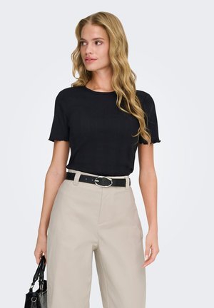 Blonde woman with wavy hair wearing black short-sleeve top, beige high-waist pants, black belt, and holding a black handbag, facing left.