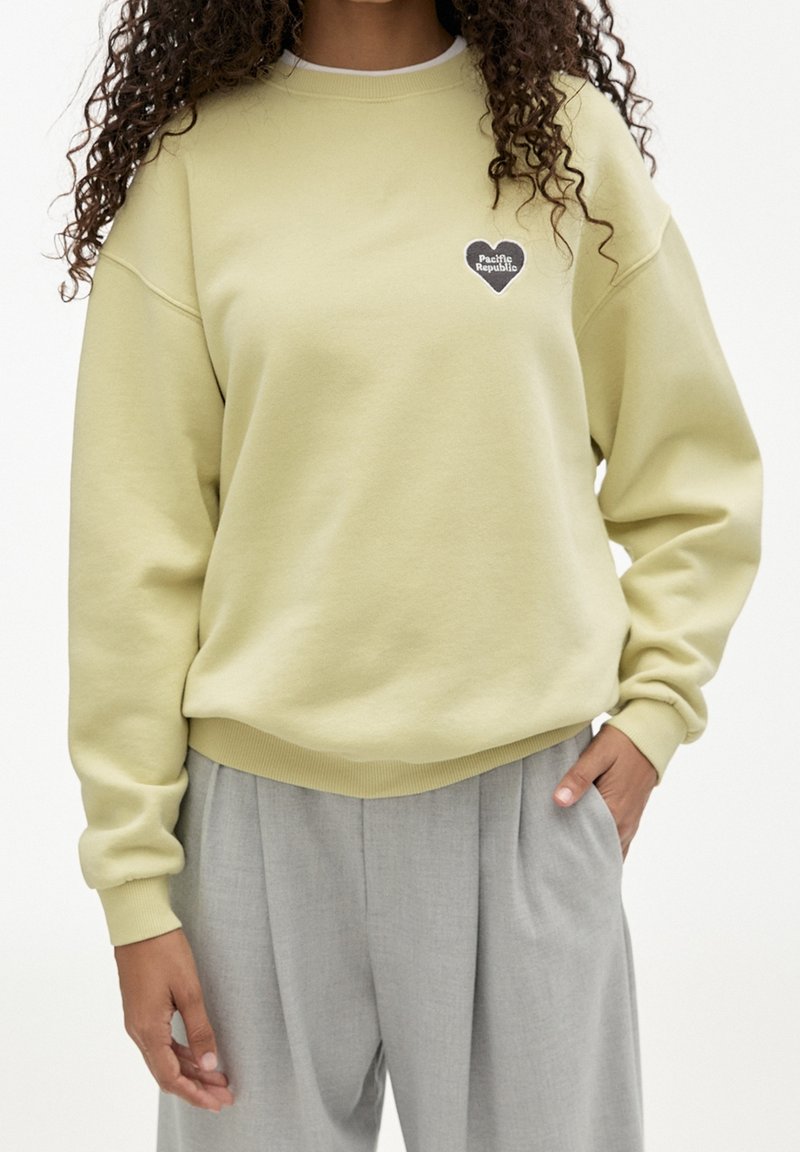 Personne portant un sweat-shirt jaune clair avec le logo en forme de cœur « Pacific Republic » sur la poitrine et un pantalon plissé gris clair, main dans la poche.