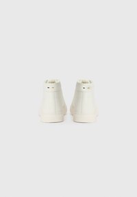Zapatillas de cuero blanco de altura media con una textura suave, que presentan acentos minimalistas en negro y dorado en el talón, sobre un fondo liso.