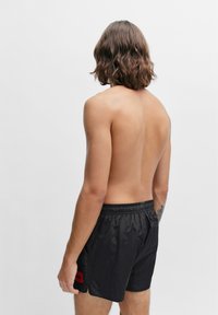 Schwarze Schwimmshorts aus leichtem Material, mit elastischem Bund und einem roten Logopatch an der Seite. Strukturiert mit glänzender Oberfläche.