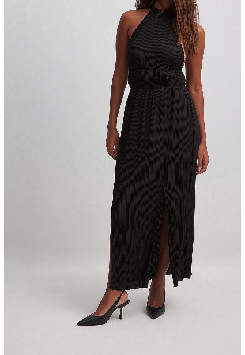 Robe maxi noire plissée à col licou avec une taille cintrée et une fente latérale. Portée avec des talons pointus noirs, mettant en valeur la texture lisse du tissu.