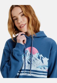 Sudadera con capucha azul que presenta un gráfico de montañas y un atardecer en tonos rosa y blanco, con tela texturizada y cordones ajustables.