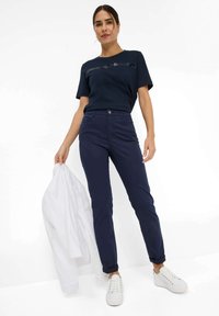 Marineblauw T-shirt met paillettenstreep, gecombineerd met marineblauwe tailored pants en witte sneakers. Witte shirt in de hand, minimalistisch ontwerp.