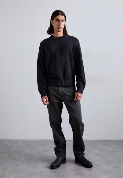 Black knitted sweater with ribbed hem, paired with dark gray jeans and black ankle boots. The design features a subtle front logo.