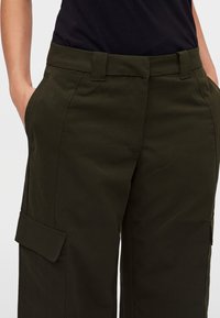 Personne portant un pantalon cargo vert foncé avec poches à rabat et passants pour ceinture, mains posées dans les poches avant, associé à un haut noir.