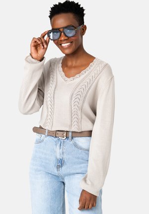 Uśmiechnięta osoba z krótkimi włosami, poprawiająca duże przyciemniane okulary, ubrana w beżowy sweter w wzory, wpuszczony w jasnoniebieskie dżinsy z brązowym paskiem.