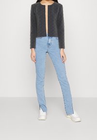 Chaqueta corta negra con un patrón texturizado y parte delantera abierta. Combinada con jeans de tiro alto en azul claro y zapatillas deportivas blancas.