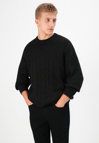 Maglione nero a treccia con scollatura tonda e maniche larghe, abbinato a pantaloni neri. La trama è spessa e accogliente.