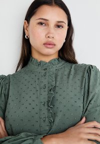 Camicia verde con bottoni, caratterizzata da pois testurizzati, colletto volant e maniche a sbuffo. Il tessuto sembra leggero e morbido.
