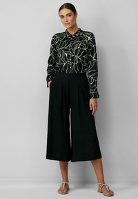 Zwarte blouse met bloemenpatroon en lange mouwen, gecombineerd met zwarte wijde broek; gedragen met lichtgekleurde sandalen.