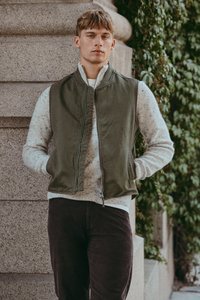 Nuori mies, jolla on päällä oliivinvihreä liivi ja kirjavavalkoinen neulepaita, seisoo kiviseinää vasten, jonka sivussa kasvaa vihreää murattia.
