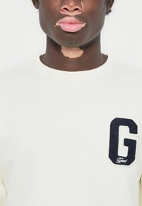 Witte sweatshirt met geribde halslijn, met een donkerblauwe, textuur "G" logo op de linkerkant van de borst. De stof lijkt zacht en comfortabel te zijn.