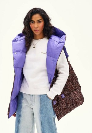 Kobieta w białej bluzie, jasnoniebieskich dżinsach i fioletowej pikowanej kamizelce bez rękawów z lampasami w panterkę, stojąca z jedną ręką w kieszeni.