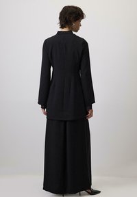 Duga crna haljina s dugim rukavima, visokim ovratnikom i lepršavom suknjom. Ima ravni kroj i suptilnu teksturiranu tkaninu.