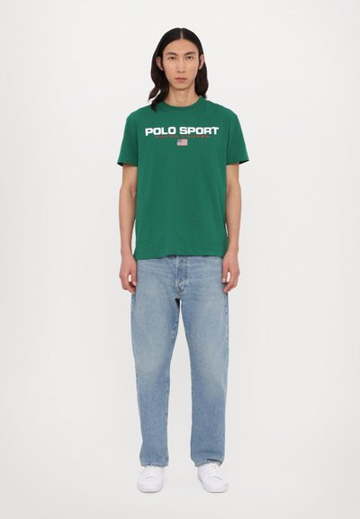 Camiseta de algodón verde con el texto "POLO SPORT" en blanco y rojo, combinada con vaqueros de un azul claro y zapatillas blancas. Diseño simple, ajuste relajado.