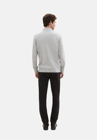 Grijze trui met lange mouwen, geribbelde manchetten en onderrand, gecombineerd met zwarte broek en zwarte schoenen, gezien van achteren.