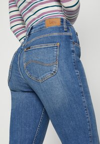 Jeans de mezclilla azul con un corte ajustado, que presentan un parche de marca de cuero en la cintura, bolsillos traseros curvados y detalles de costura amarilla.