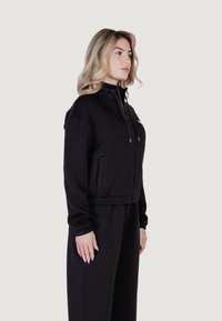 Juodas zip-up švarkas su aukštu apykakliu, dviem priekinėmis kišenėmis ir elastinėmis rankovėmis. Pagamintas iš lengvos medžiagos, sukurtas prigludusiam siluetui.