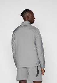Pilkas lengvas švarkas su aukštu kaklu ir ilgomis rankovėmis, turintis tekstūruotą audinį ir juodą Reebok logotipą ant nugaros.