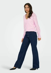 Cárdigan de punto color rosa claro con escote en V y patrón de diamantes, combinado con pantalones vaqueros de pierna ancha de color azul oscuro y zapatos de tacón negro de punta.