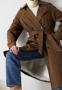 Bruine trenchcoat met een ceintuur in de taille, dubbele knopen en een zachte, crèmekleurige voering. Model draagt een blauwe spijkerbroek eronder.
