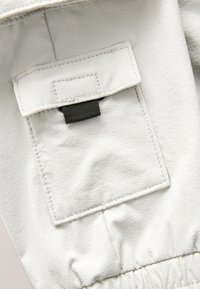 Tasca in tessuto grigio chiaro con patta fissata da linguetta nera, cucita su capo abbinato con orlo elastico sotto.