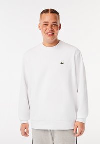 Weißes Sweatshirt mit langen Ärmeln, gerippten Bündchen und Rundhalsausschnitt. Verfügt über ein kleines grünes Logo auf der Brust. Glatte Textur.