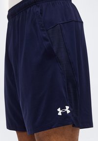 Ναυτικό μπλε αθλητικό σορτς με ελαστική μέση, πλαϊνές διχτυωτές πινακίδες και λευκό λογότυπο Under Armour στο αριστερό πόδι, φορεμένο από άτομο.