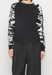 Svart tröja med ärmar i blommönster i vitt och grått. Designen inkluderar en rund halsringning och ribbad mudd, tillsammans med plisserade svarta byxor.