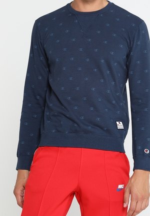 Man wearing a navy blue Champion sweatshirt with logo pattern and red Nike pants with logo on left thigh against a plain background.