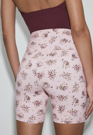 Woman wearing high-waisted pink biker shorts with small purple floral print and a dark maroon sleeveless top, shown from the back.