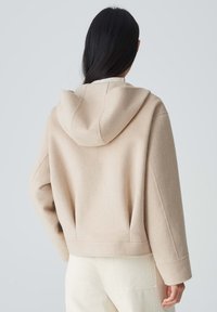Beige Wollmischung-Hoodie mit einer weichen Textur, ausgestattet mit einer Fronttasche, weiten Ärmeln und einem lockeren Schnitt, von hinten gezeigt.