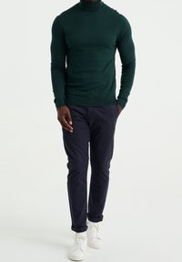 Mörkgrön polotröja kombinerad med marinblå slim fit-byxor och vita sneakers, med en figurnära design och ribbad textur vid kanterna.