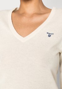 Pull crème en V fabriqué dans un doux tricot. Présente un petit logo GANT bleu sur la partie gauche de la poitrine. Design simple avec des bords côtelés.