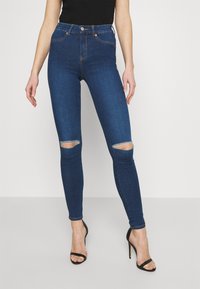 Jeans skinny de tiro alto, de color azul oscuro, con desgastes en las rodillas y costuras en naranja, presentando un cierre clásico de botón. Textura de denim suave.