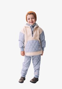 Helles blaues, gestepptes Hoodie mit hellen Fleece-Akzenten und einer Vorderfronttasche, kombiniert mit passenden gesteppten Hosen. Braune Mütze und dunkle Stiefel.