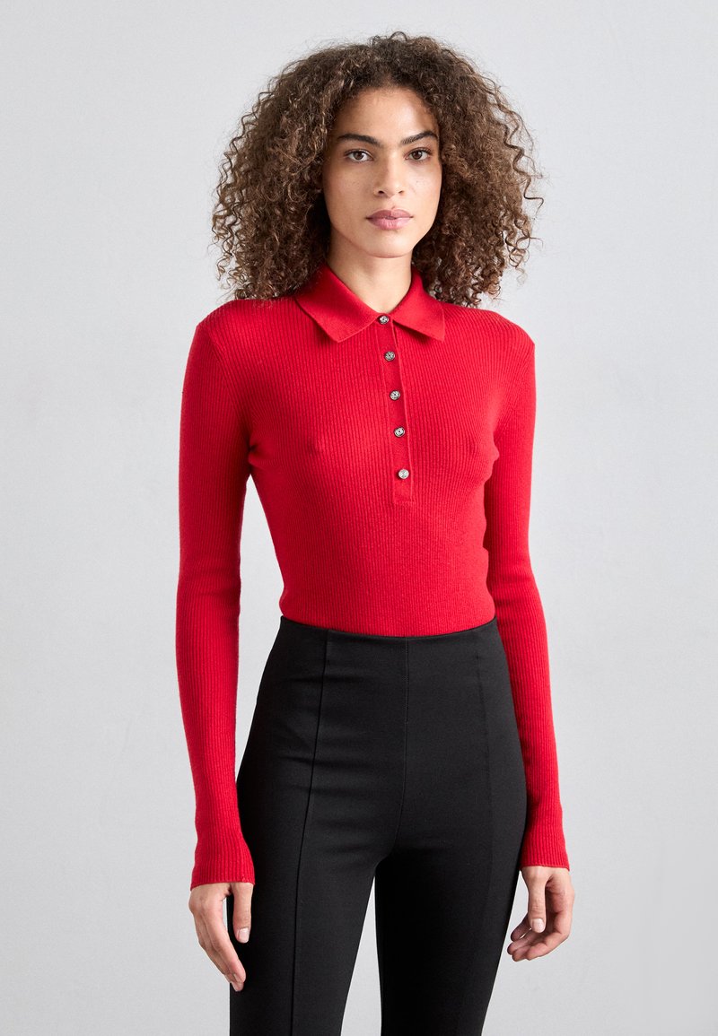 Röd ribbad långärmad polo med krage och sex metallknappar. I kombination med högmidjade svarta byxor. Slät textur.