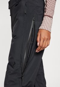 Black ski pants feature zippered side pockets and a sleek design. Textured fabric with a slight sheen and embossed logo detail.