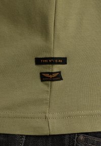 Tela de camiseta color verde olivo con una textura suave. Presenta dos etiquetas negras cosidas, una etiquetada "TIPO N°: C-46" y la otra "CLÁSICO AMERICANO."