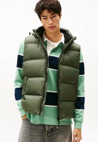 Chaleco tipo puffer en verde oliva con cierre de cremallera, con acolchado tipo colcha. Usado sobre una camiseta de rayas en tonos verdes y azul marino de manga larga.