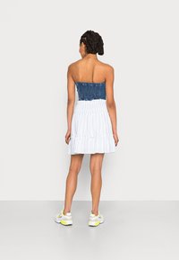 Vestido sem alças com um corpete de ganga e saia branca em camadas. Apresenta um design franzido no corpete, tecido texturizado e bainha com folhos.