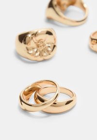 Guldringe sæt, der omfatter to glatte ringe, der er sammenflettet, og tre ekstra statementsringe med blomsterdetaljer. Poleret metallisk finish.