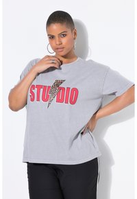 Camiseta gris de gran tamaño con el texto "STUDIO" en rojo intenso y un diseño de rayo de estampado de leopardo. Tejido suave de algodón con mangas cortas.
